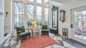 Una habitación amplia con ventanas grandes, un tapete estampado, dos sillones verdes, una mesita de madera, un espejo y un reloj.