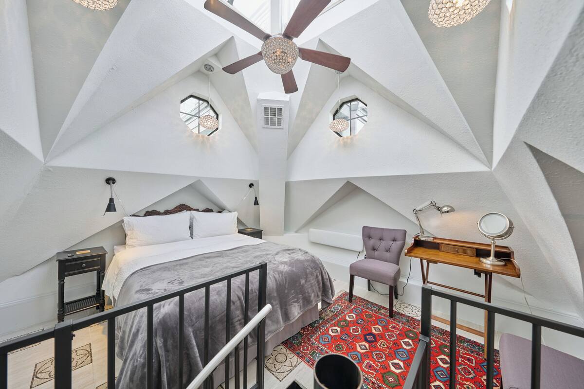 A modern bedroom with a sloped ceiling, a bed, a chair, a desk with a mirror, and a red patterned rug.