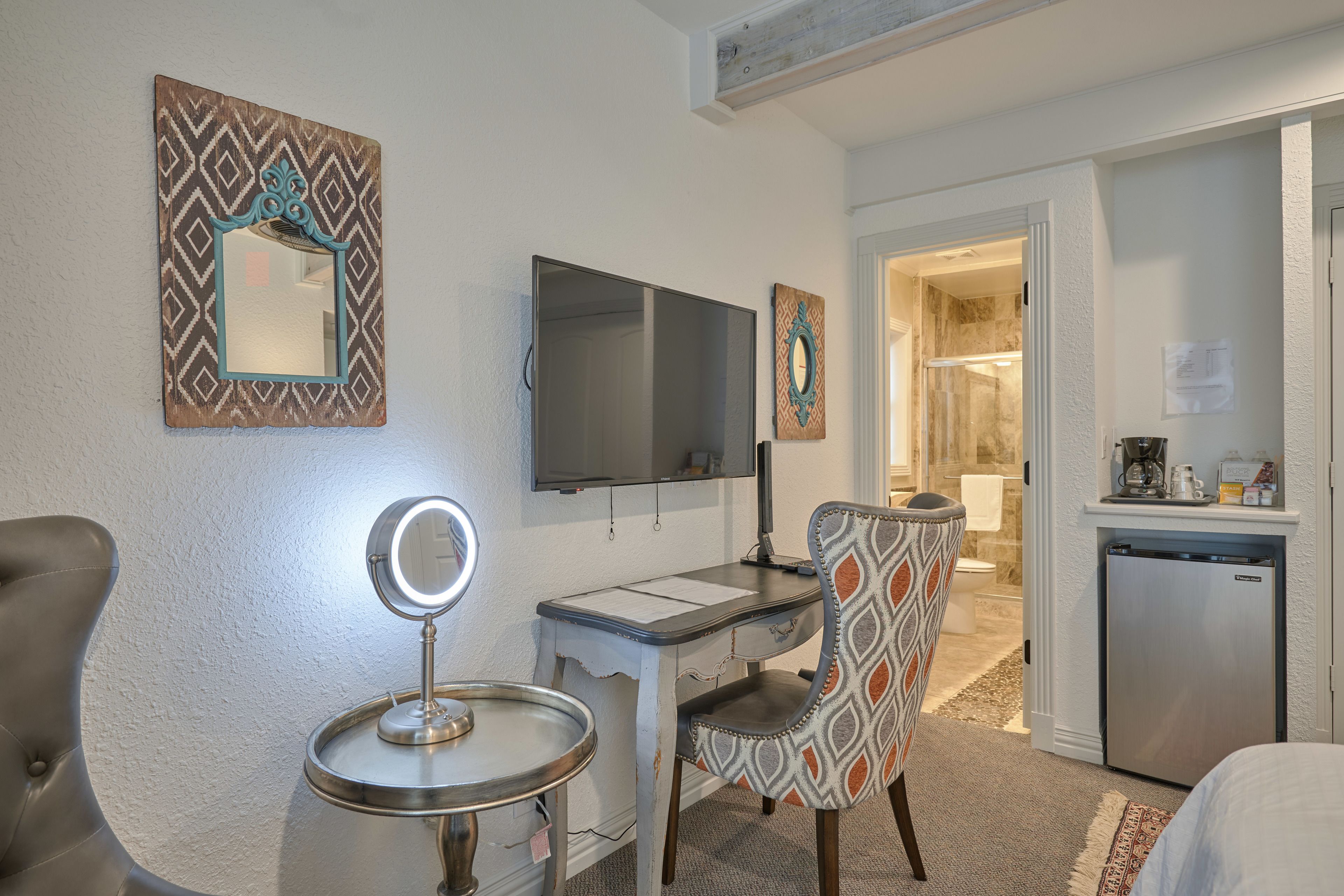 A hotel room with a desk, chair, TV, and a small kitchenette.