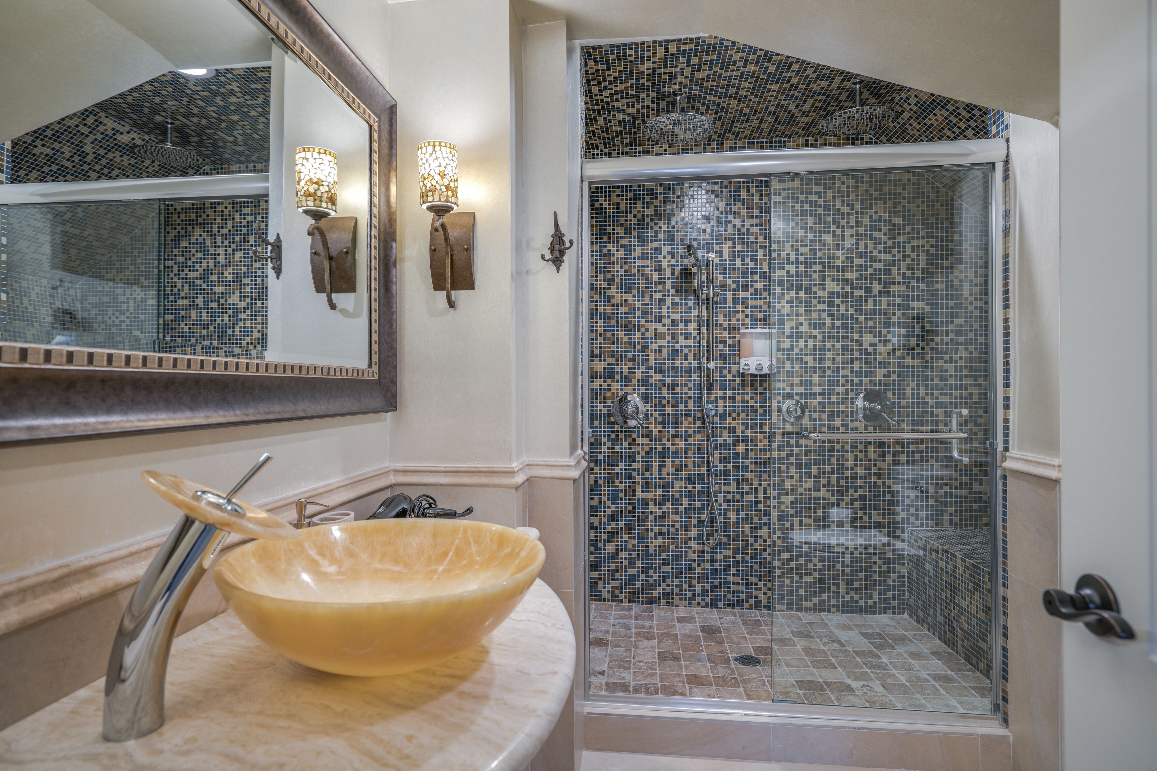 A bathroom with a large bowl sink, a glass-enclosed shower with mosaic tiles, and wall-mounted sconces.