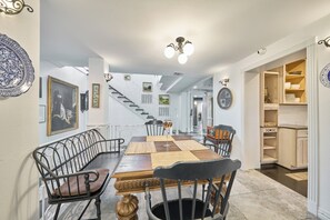 Un comedor con una mesa de madera, sillas de metal negro y un mantel tejido. Se ve una escalera al fondo que conduce a otra habitación.