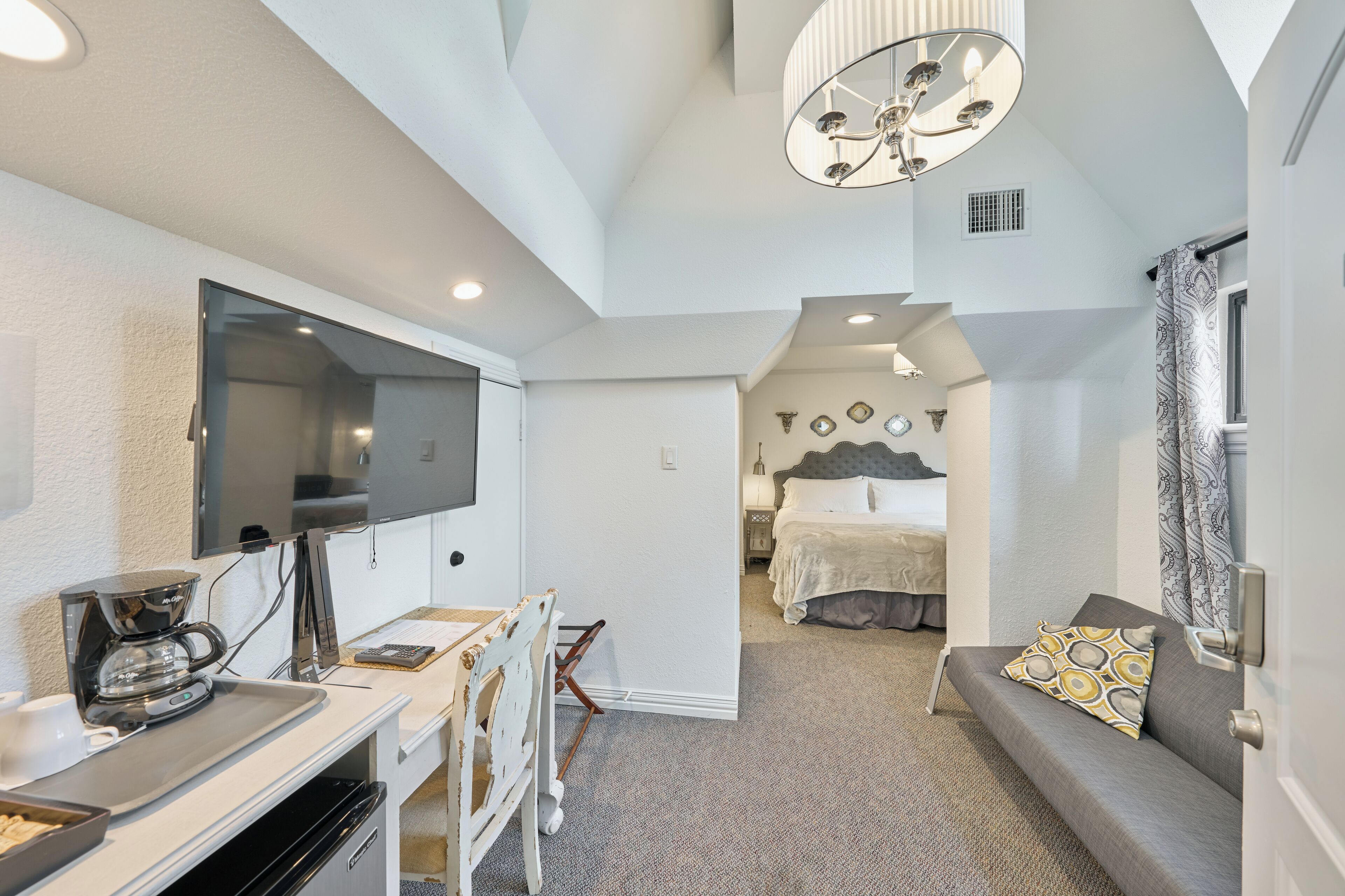 A modern hotel room with a flat-screen TV, a coffee maker, a desk with a chair, a sofa with a patterned pillow, and a bed with a headboard.