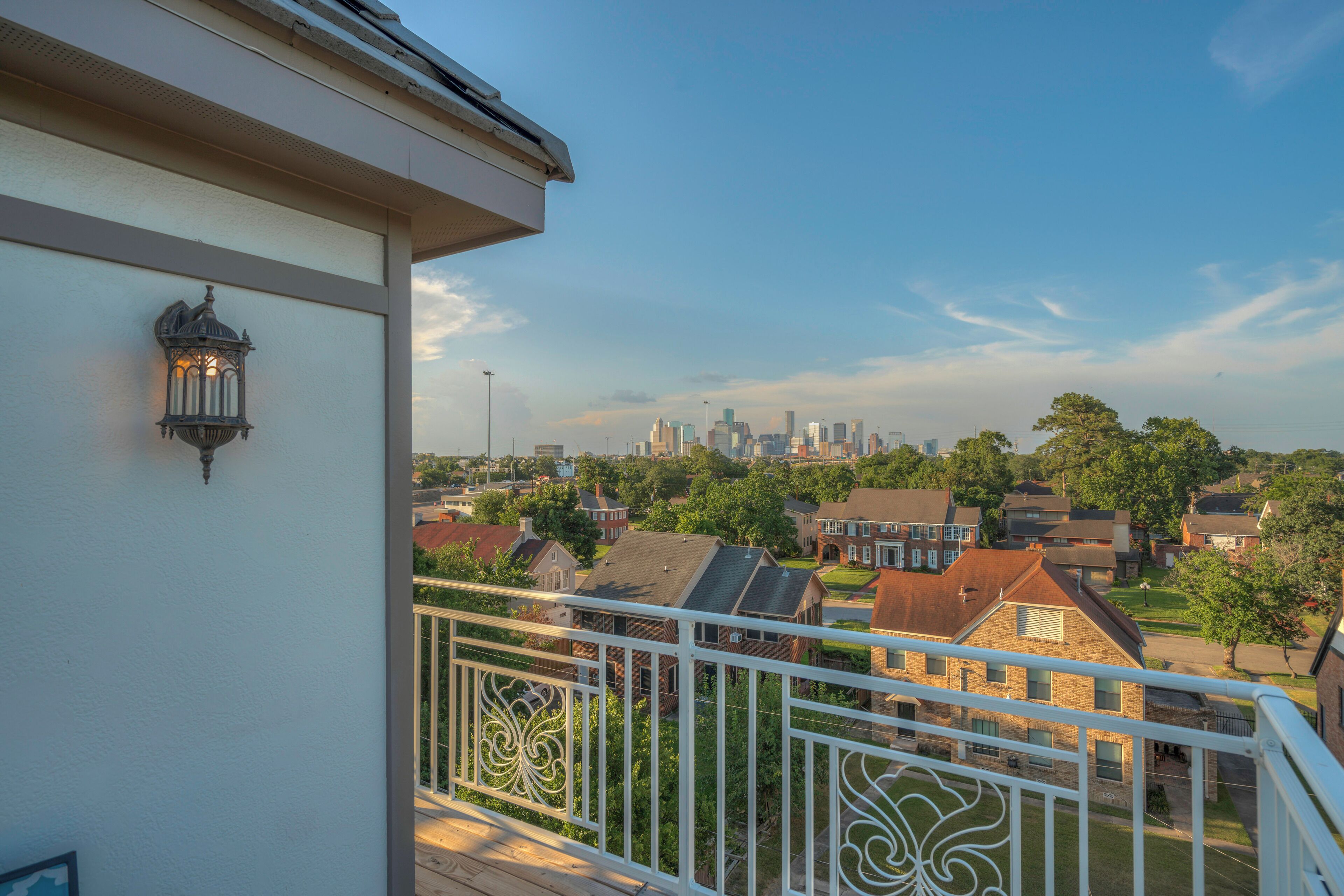 A balcony with a railing, a wall-mounted light fixture, and a view of a residential neighborhood with houses and a distant city skyline.