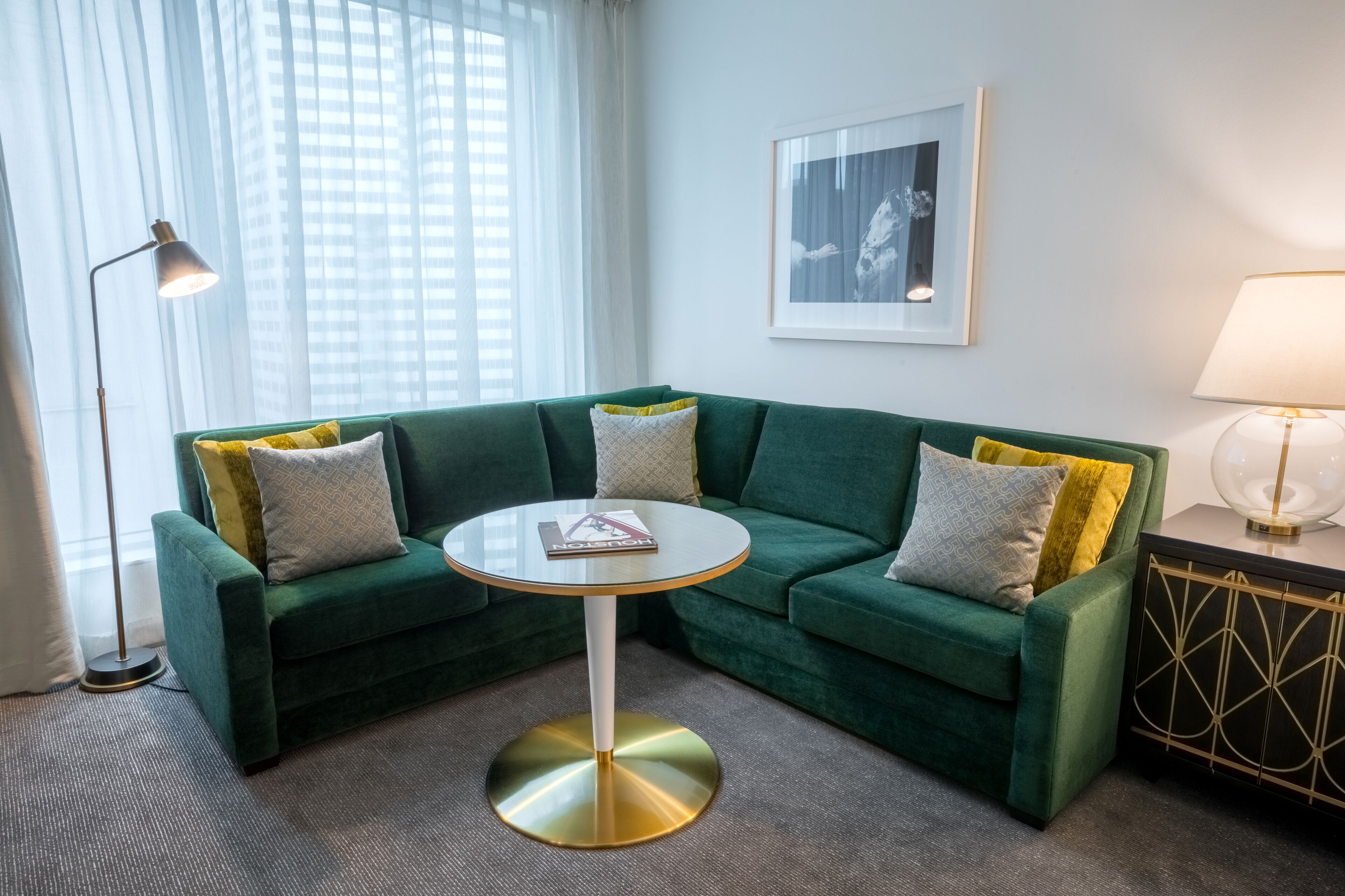 Um sofá verde com almofadas amarelas, uma mesa redonda com base dourada e um quadro na parede.