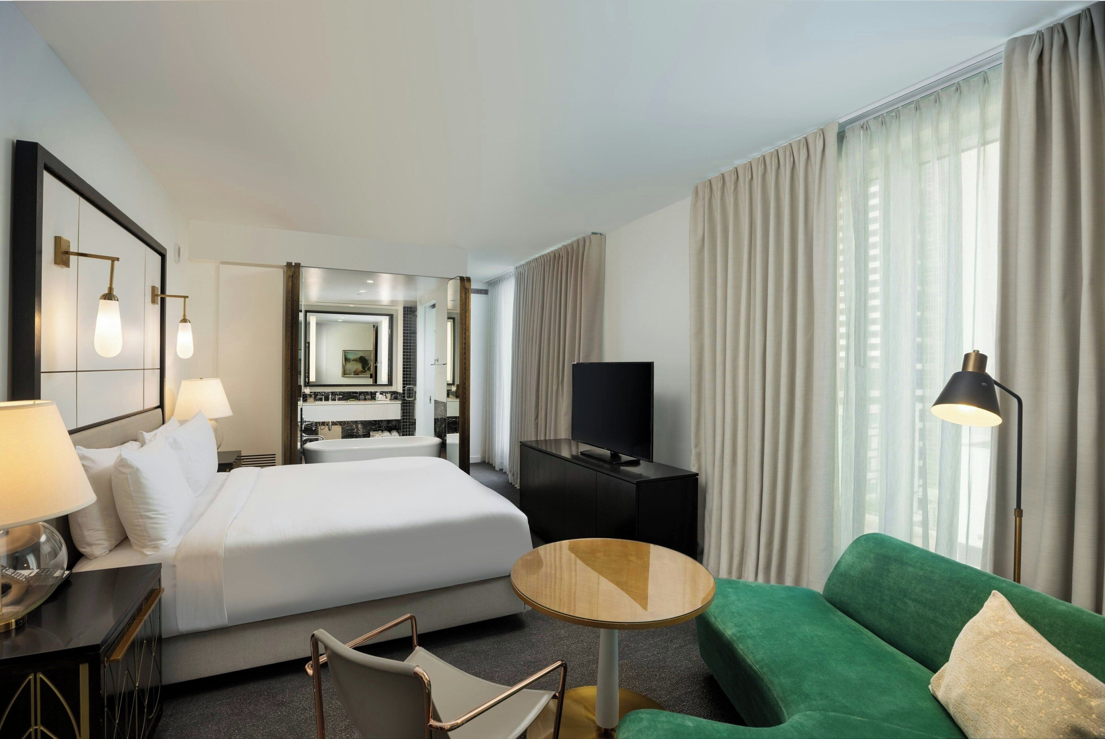A hotel room with a bed, a green sofa, a small round table, a TV, and a mirror.
