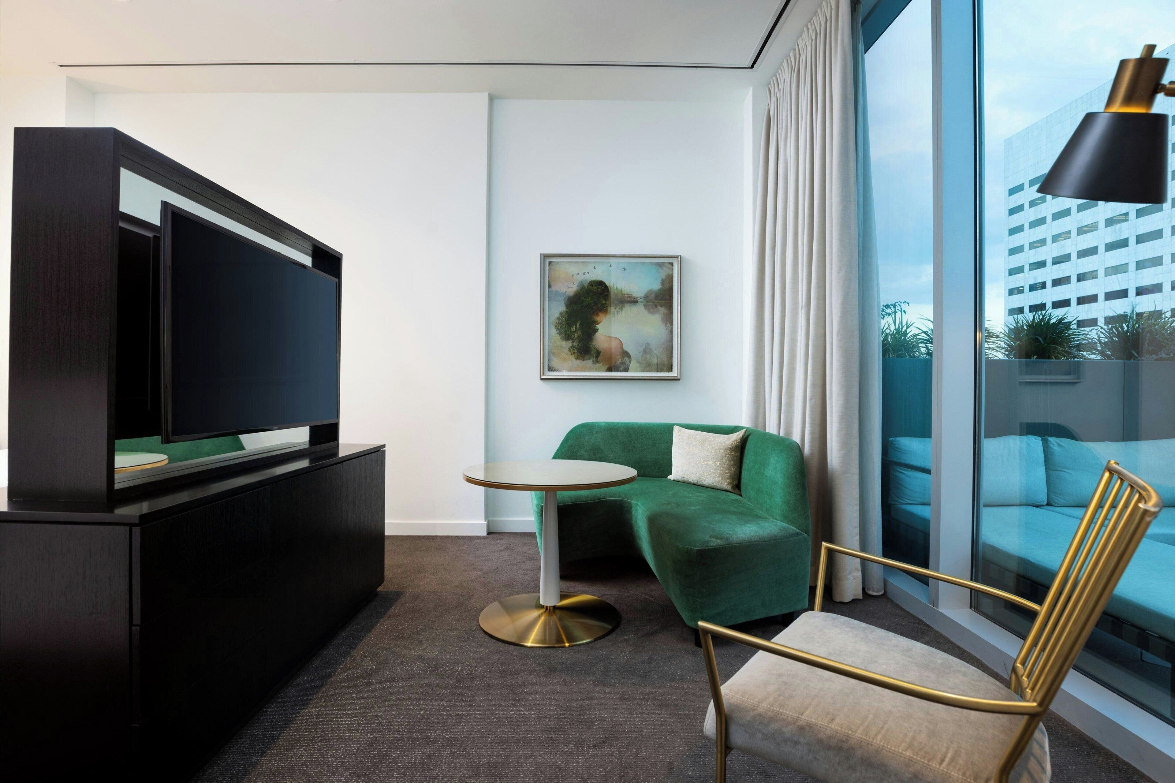 A modern hotel room with a flat-screen TV, a green sofa, a round table, and a gold-framed chair.