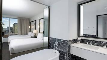 A hotel room with two beds, a large window with a city view, a bathroom with a bathtub, and a vanity with a mirror.