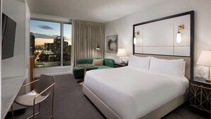 A hotel room with a large bed, a desk with a chair, a green sofa, and a view of a cityscape.