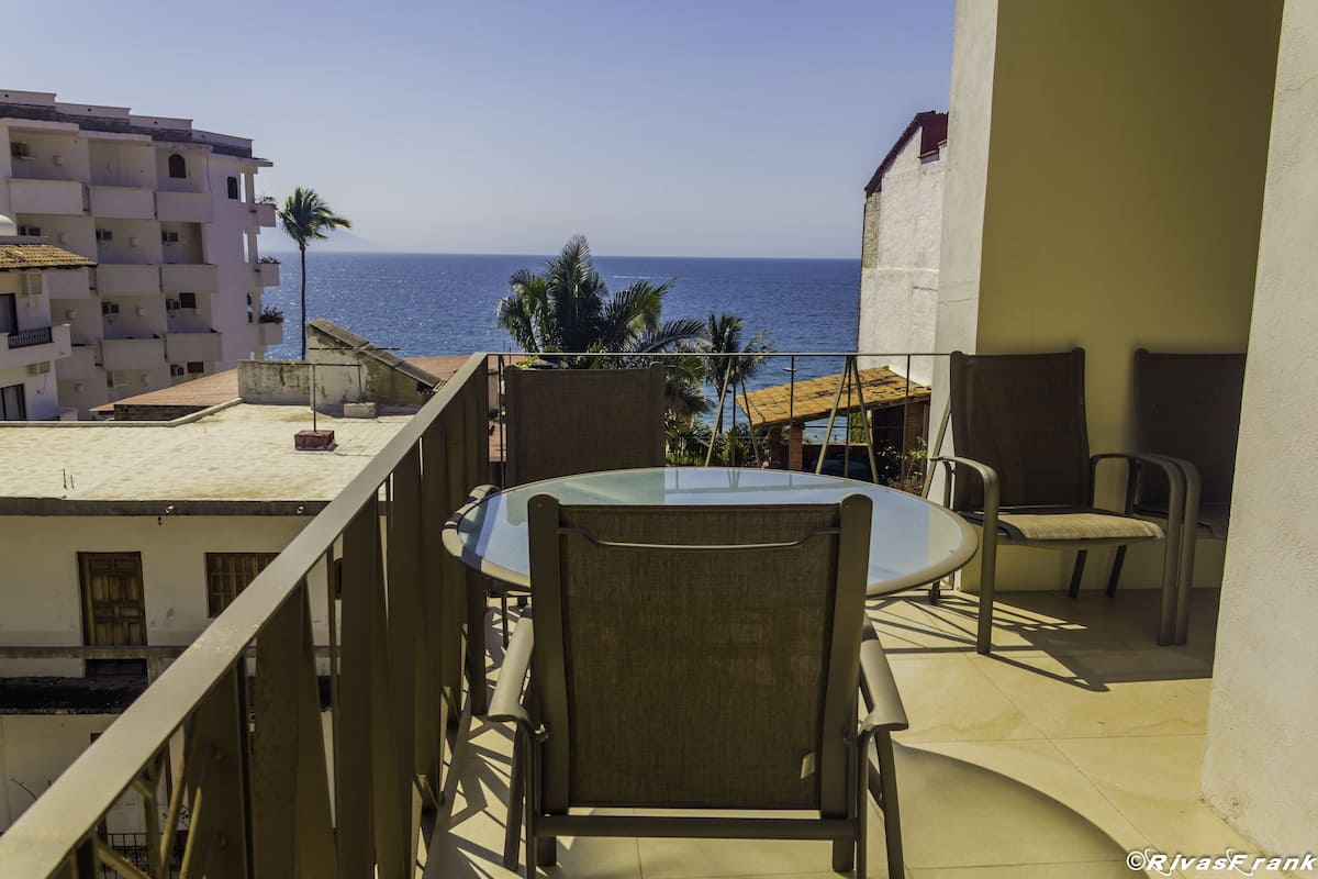 Un balcón con una mesa y sillas de vidrio, con vista a una playa y palmeras.