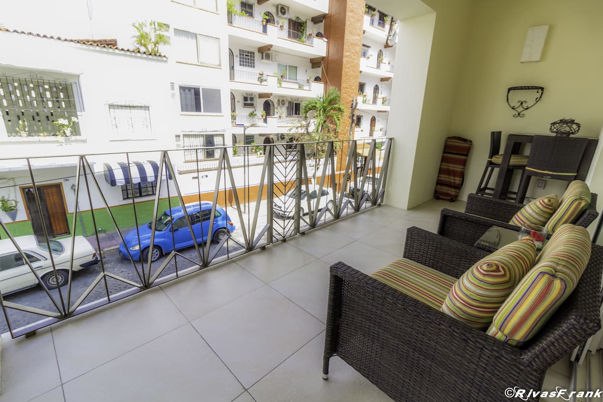 Un balcón con mobiliario de mimbre, vistas a coches estacionados y un edificio con balcones al fondo.