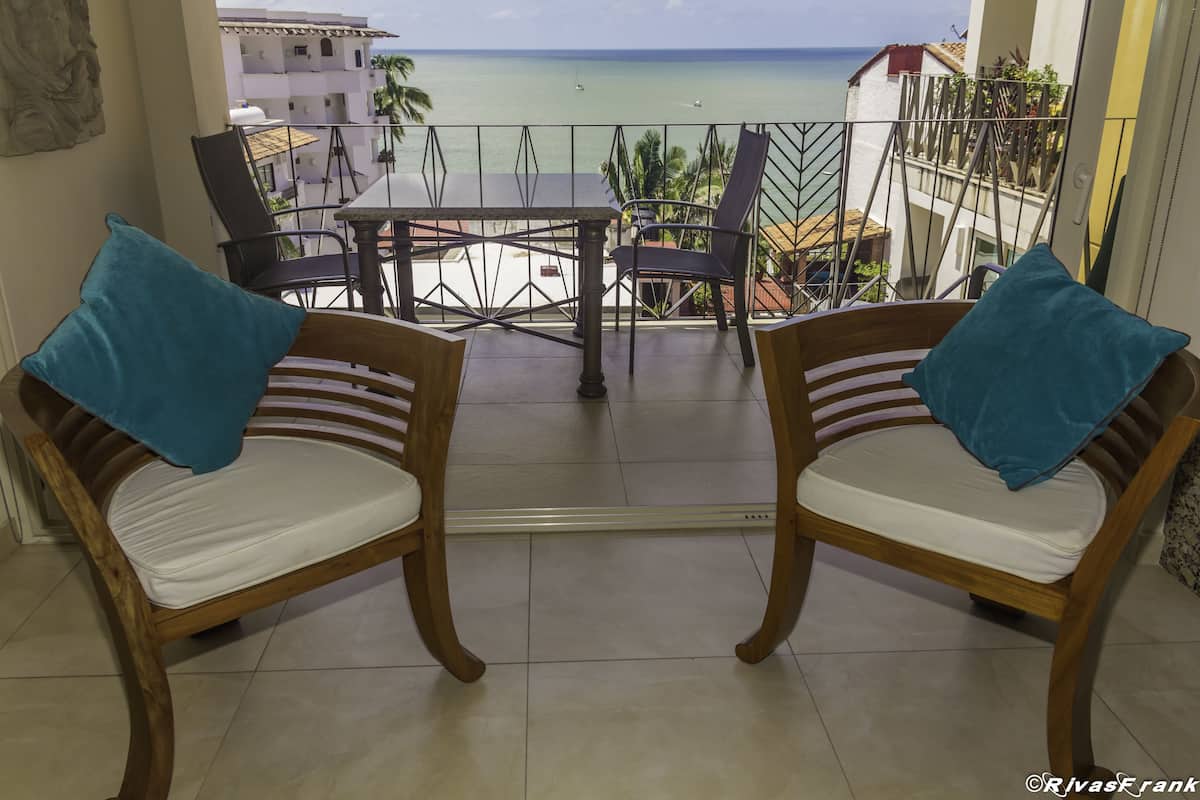 Un balcón con dos sillas de madera y una mesa, con vistas a una playa con edificios y palmeras.