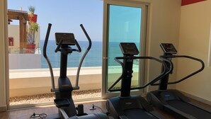 Un gimnasio en casa con dos bicicletas estáticas y una cinta de correr, con vistas a un balcón y al mar.