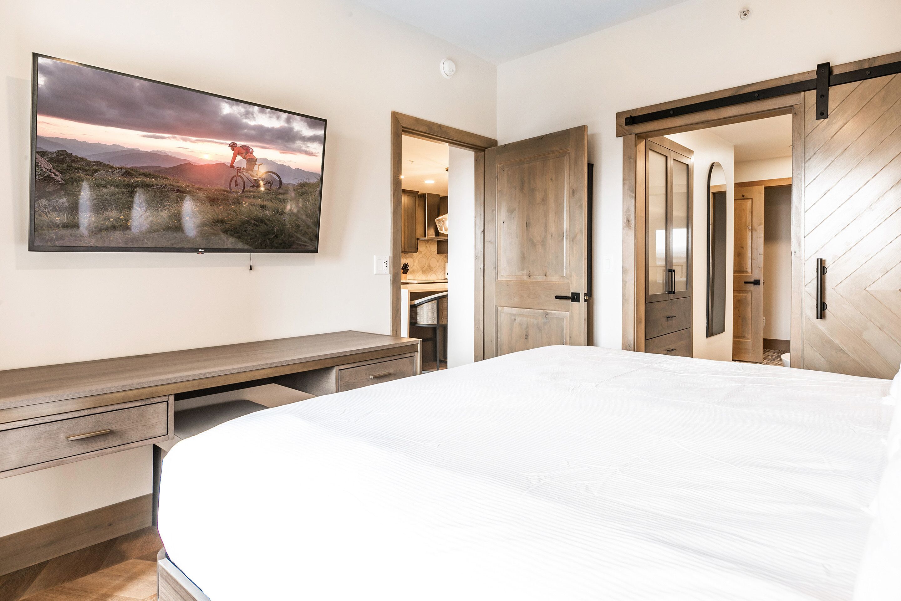 A bedroom with a large bed, a TV mounted on the wall, a wooden desk, and a sliding door leading to a bathroom.