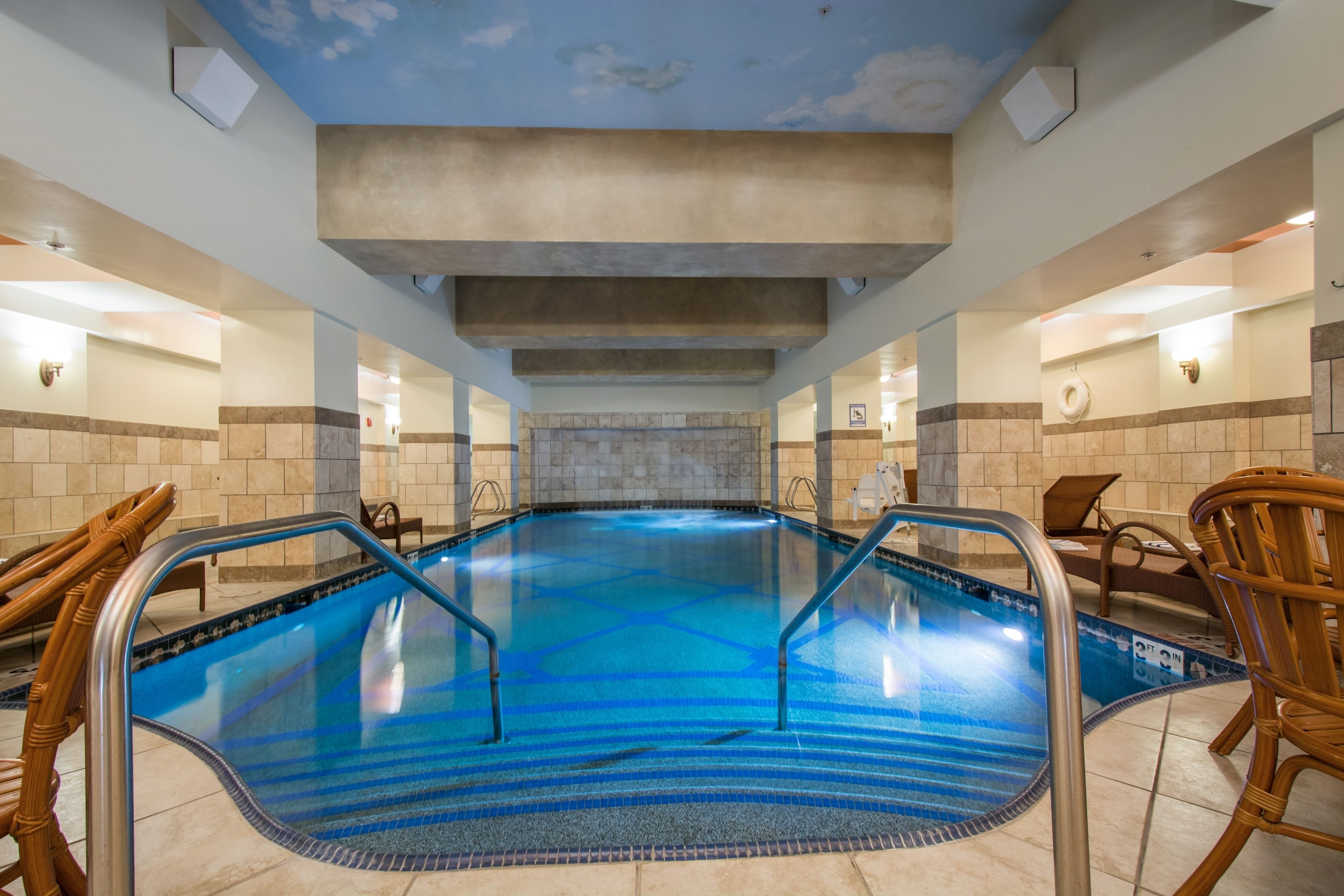 A large indoor swimming pool with clear blue water, surrounded by wooden chairs and a stone wall in the background.