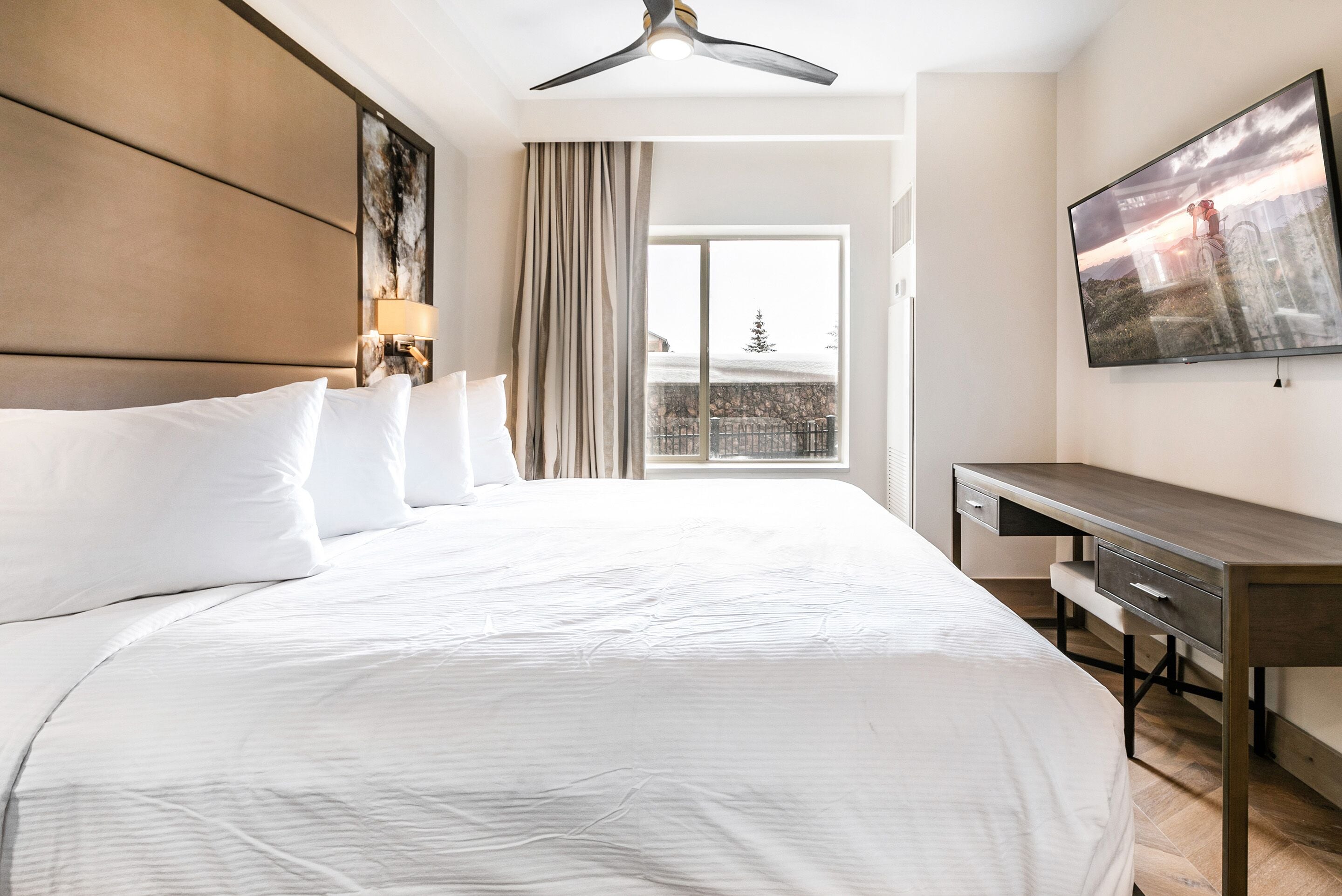Ein Hotelzimmer mit einem großen Bett, einem Schreibtisch, einem Deckenventilator, einem Fernseher und einem Fenster mit Blick auf eine verschneite Landschaft.
