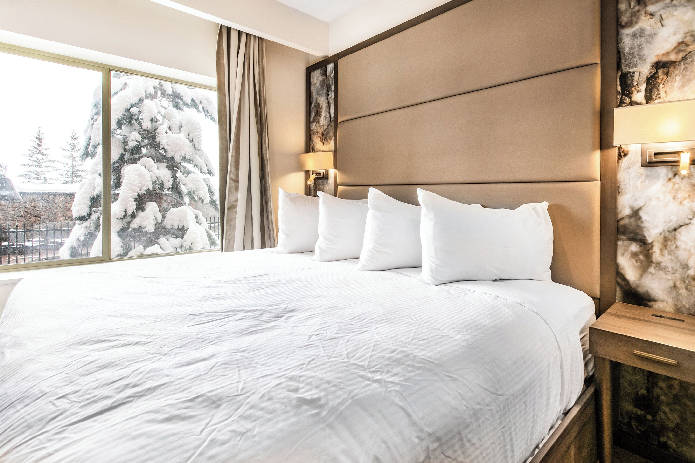 A hotel room with a large bed, a headboard with a light fixture, a window with a view of snow-covered trees, and a marble wall.