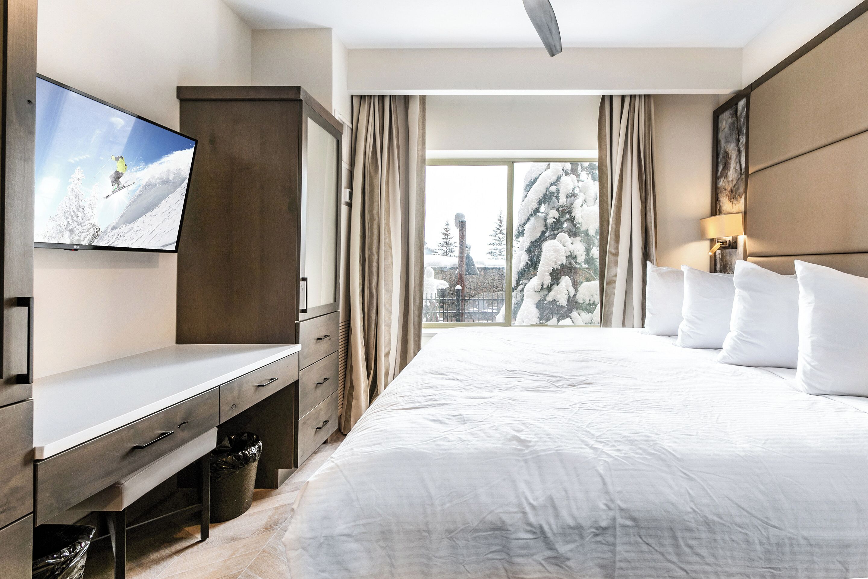 Habitación de hotel con una cama grande, un televisor montado en la pared, un escritorio con cajones y vistas a árboles cubiertos de nieve a través del ventanal.