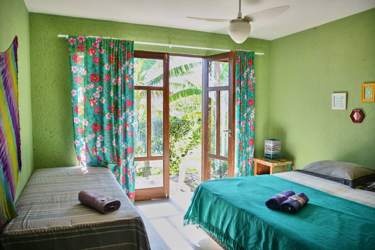 Ein Zimmer mit zwei Betten, einem Deckenventilator und Blick auf Grünfläche.