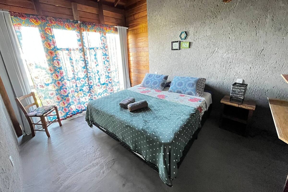 Ein Schlafzimmer mit einem Bett, einem Sessel, einem kleinen Tisch und einem Fenster mit Vorhängen.