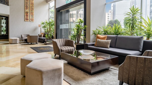 Un hall d’entrée spacieux, doté d’un canapé moderne, de fauteuils et d’une table basse en verre. On y trouve également des plantes en pot et une grande fenêtre donnant sur la ville.