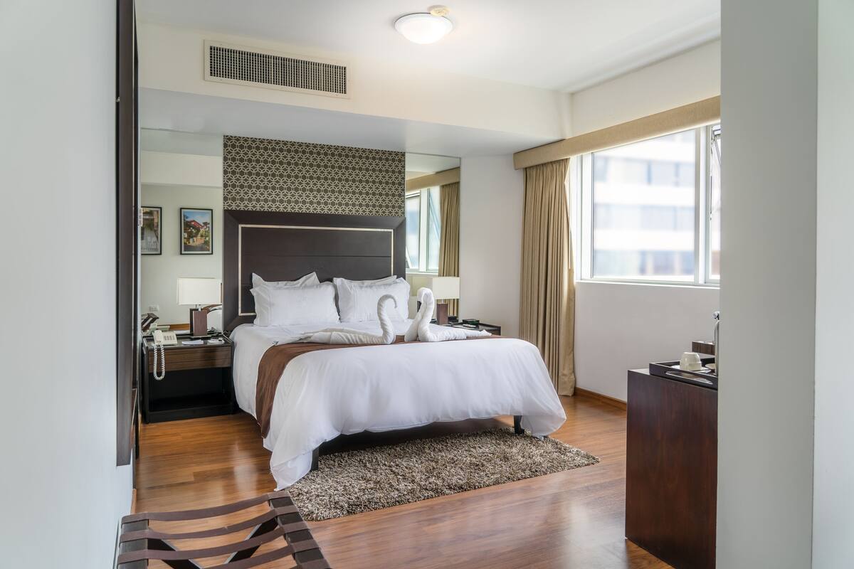 A modern hotel room with a large bed, a wooden floor, a patterned headboard, a bedside table with a lamp, and a window with curtains.