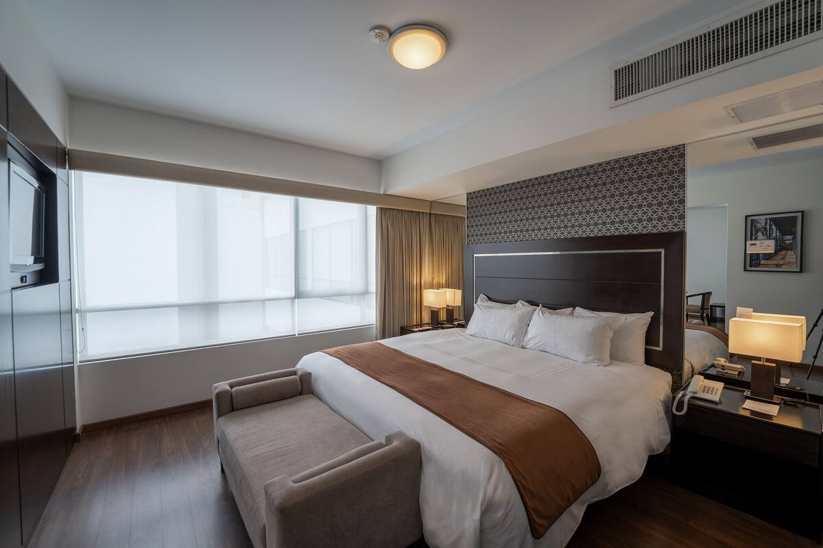 A modern hotel room with a large bed, a bedside table with a lamp, a nightstand with a phone, a window with curtains, and a wall-mounted artwork.