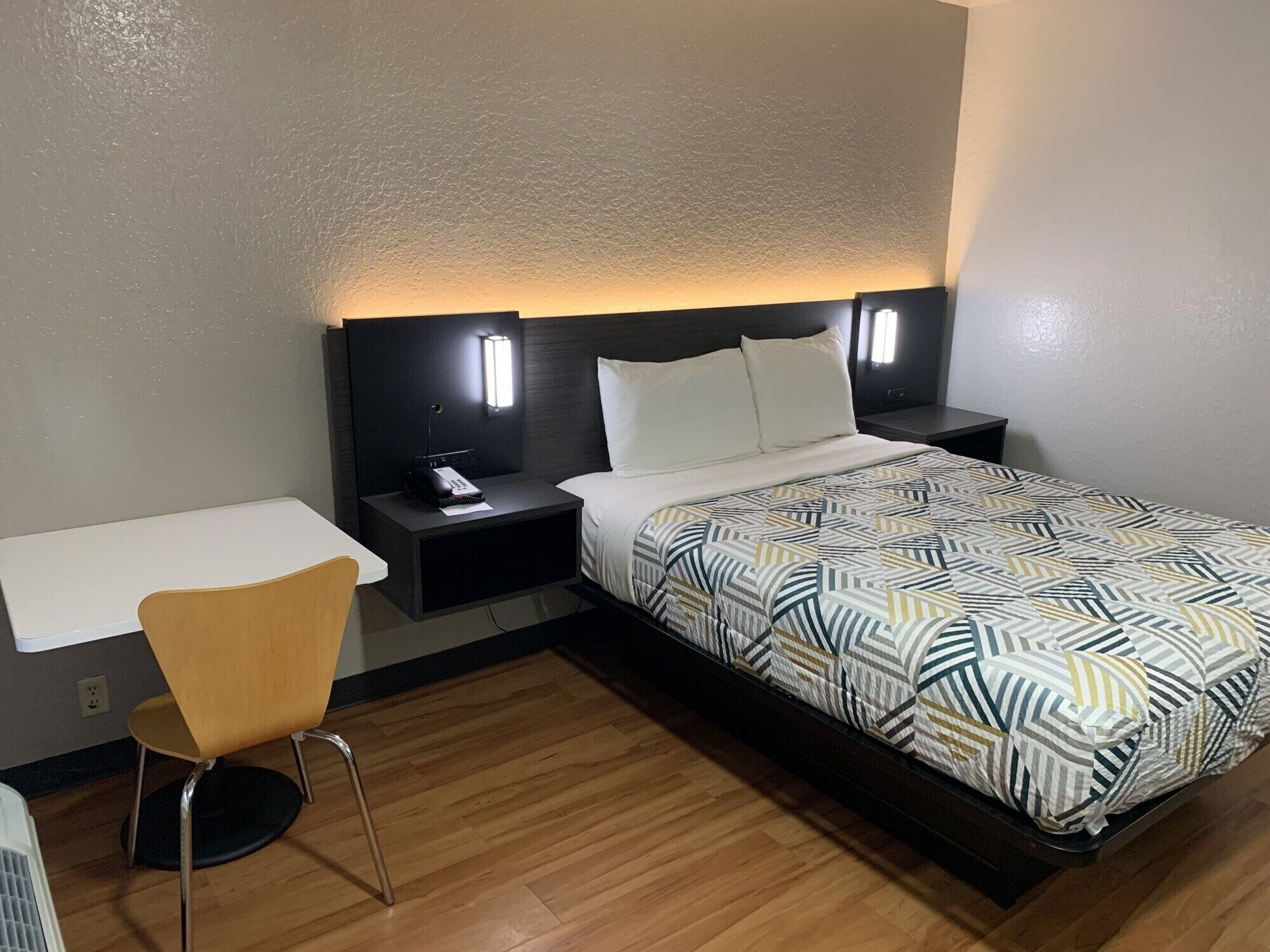 A hotel room with a bed, bedside table, chair, and a desk.