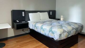 Una cama bien hecha con una colcha estampada, una mesita de noche con un teléfono y una lámpara, y un estante fijado a la pared.