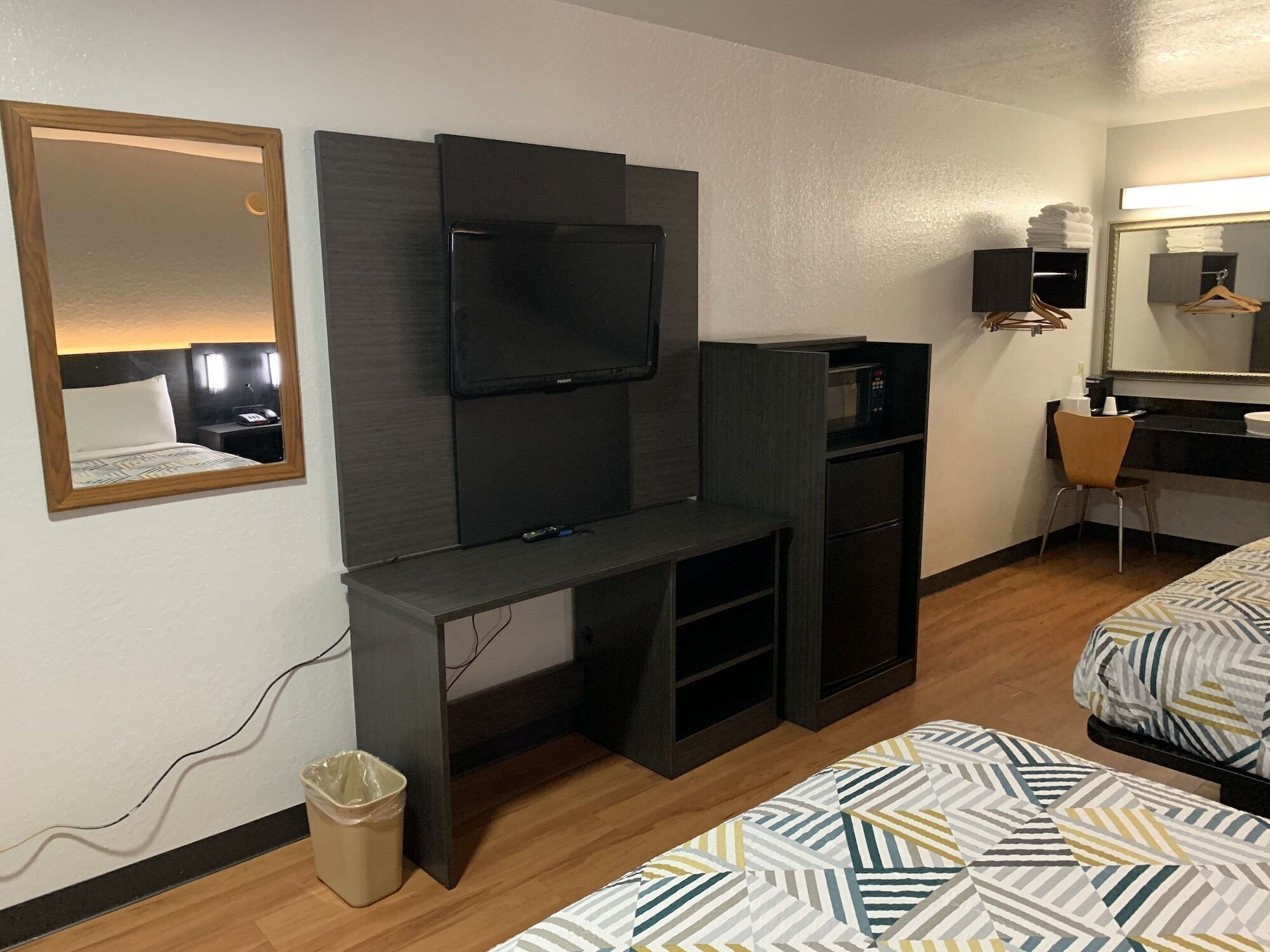 Habitación de hotel con cama, televisor montado en un panel oscuro, escritorio con silla, kitchenette pequeña y espejo.