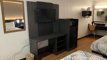 Habitación de hotel con cama, televisor montado en un panel oscuro, escritorio con silla, kitchenette pequeña y espejo.