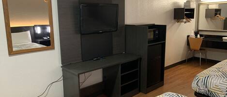 Habitación de hotel con cama, televisor montado en un panel oscuro, escritorio con silla, kitchenette pequeña y espejo.