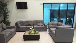 A modern waiting area with grey sofas, a central coffee table with flowers, a flat-screen TV, and a glass door leading outside.