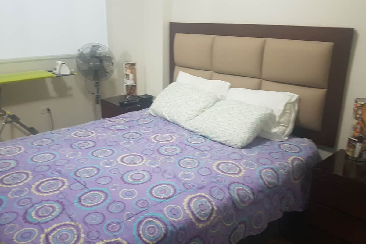 A bed with a patterned bedspread, a bedside table with a lamp, and a wall-mounted fan.