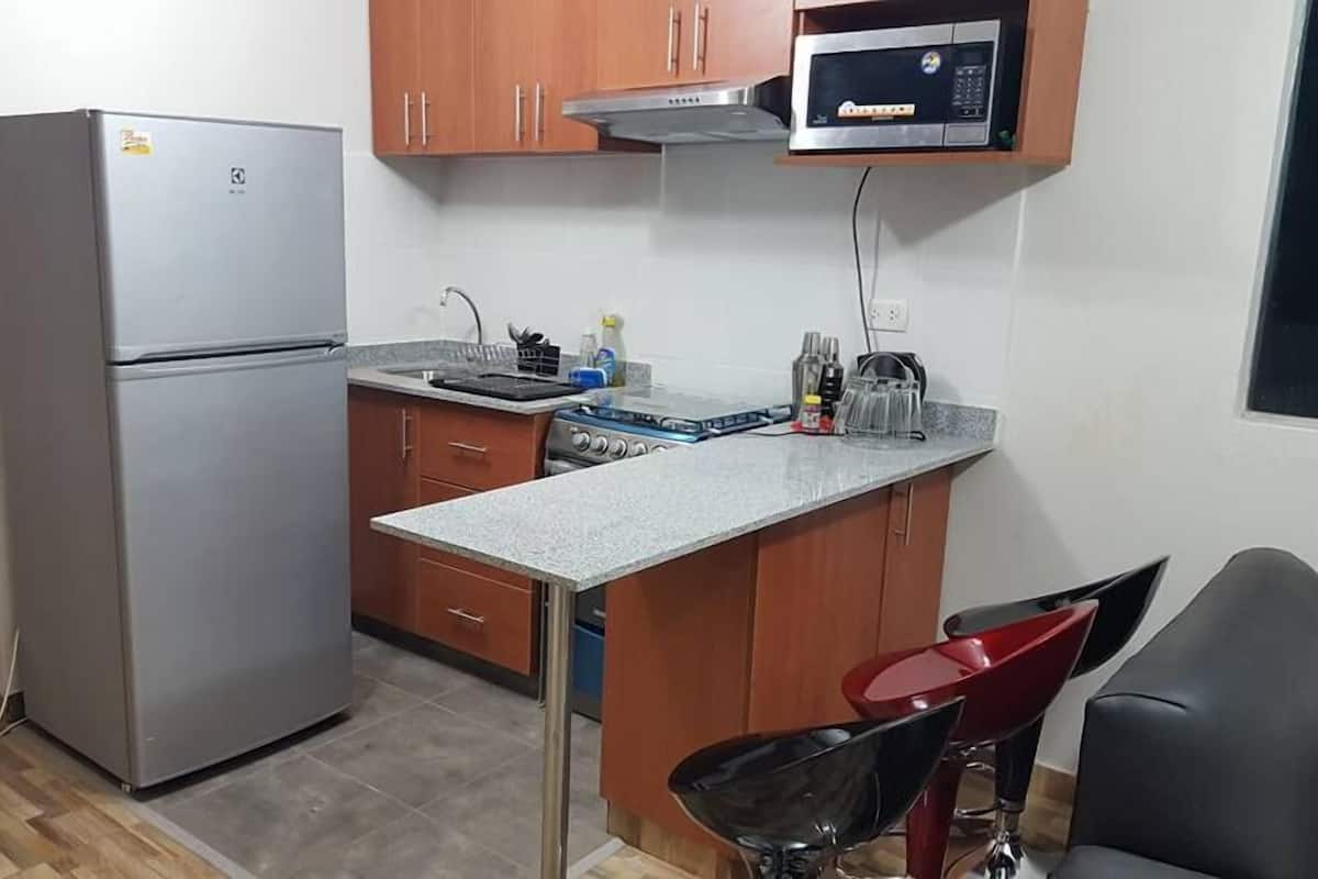 A compact kitchen with a refrigerator, microwave, stove, and a small dining area with a chair.