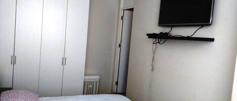 A bedroom with a bed, a wardrobe, a television mounted on the wall, and a laundry basket.