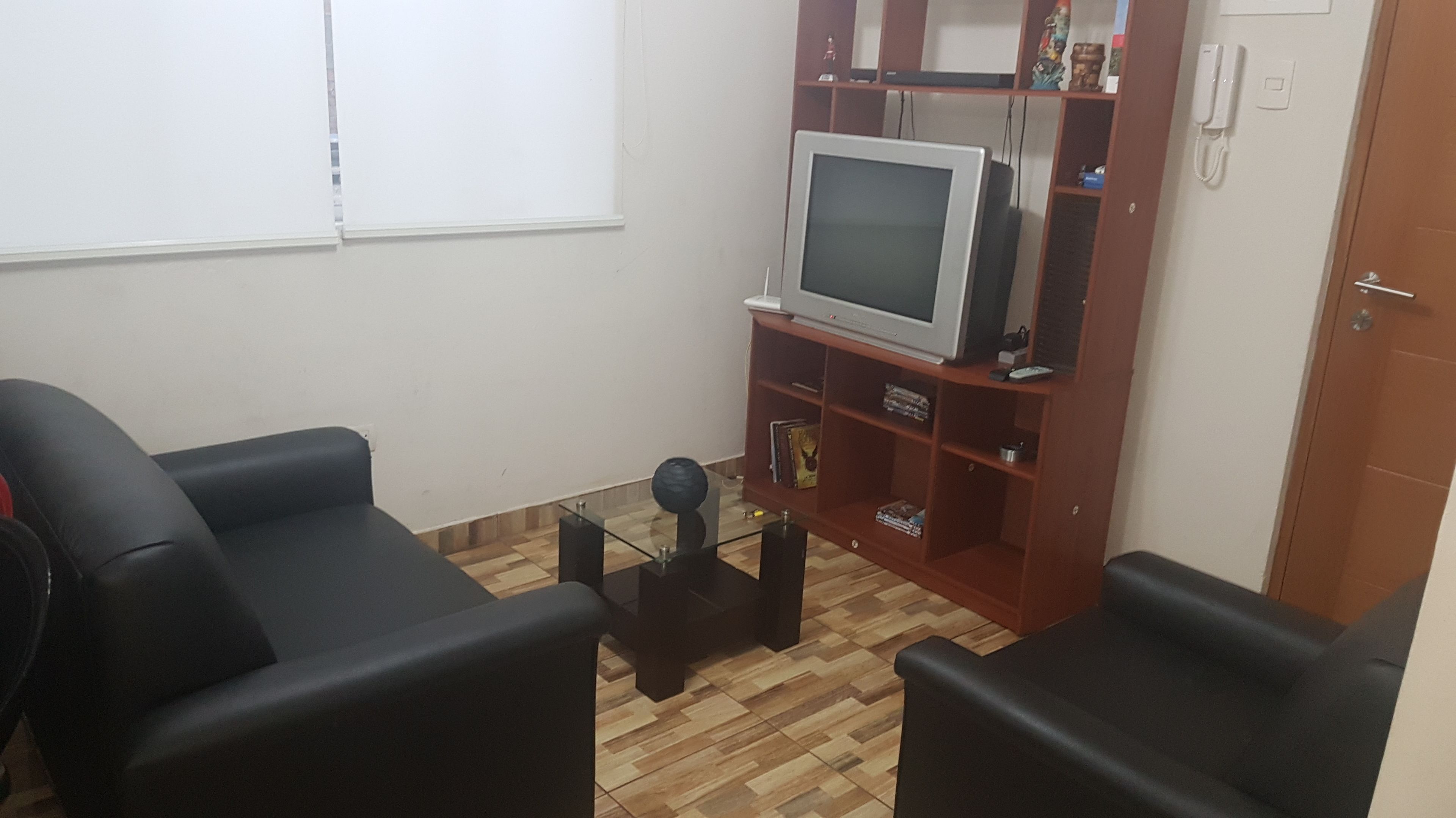 A living room with a television on a wooden stand, two black sofas, and a glass coffee table.