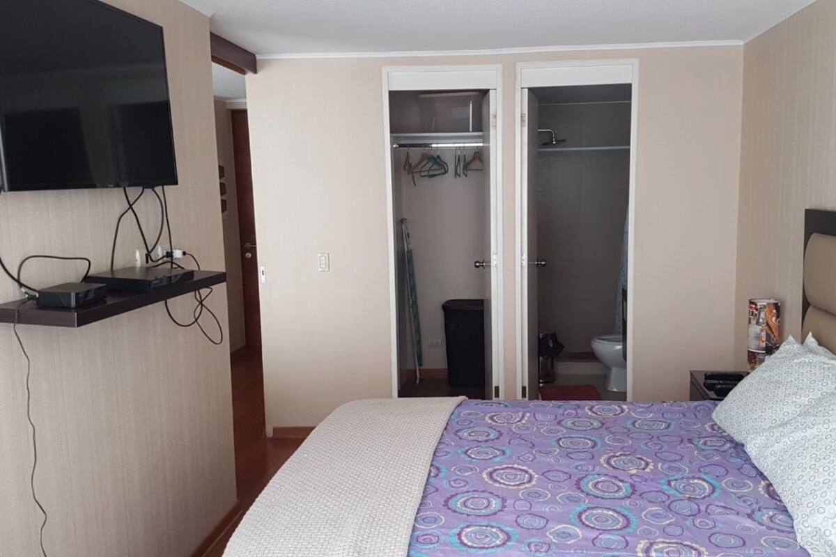 A bedroom with a bed, a TV mounted on the wall, a closet, and a bathroom visible through an open door.