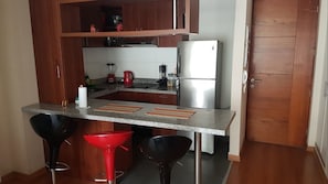 A compact kitchen with a refrigerator, a sink, and a countertop. There are two stools and a red sink.