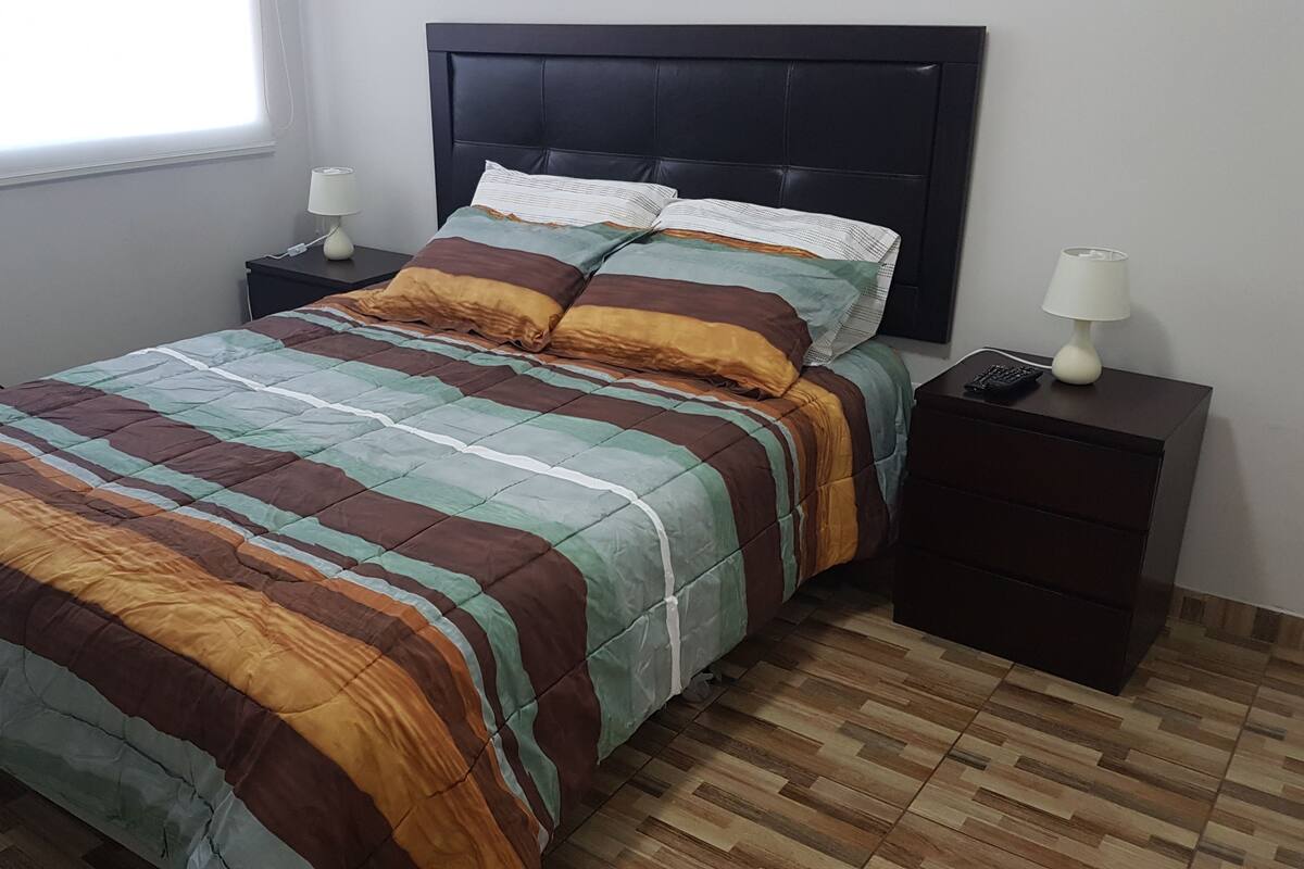 Uma cama bem arrumada com colcha listrada, duas mesas de cabeceira com abajures e uma janela com persianas.