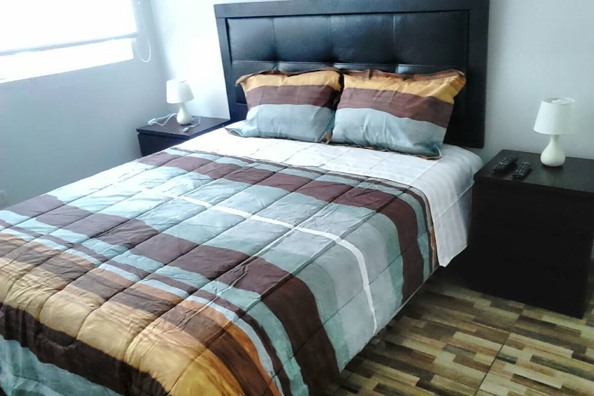 Uma cama com colcha xadrez, cabeceira escura e duas mesas de cabeceira com abajures.