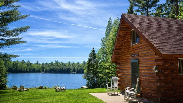 Eine Holzhütte mit Blick auf einen See, einem Picknicktisch und zwei Stühlen vor der Tür.