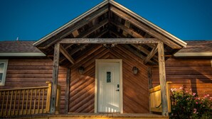 A wooden cabin with a white door, a porch with a wooden railing, and a small garden with pink flowers. - Rustic Cabin - Jetted Tubs - Wi-Fi - Wonderful Views - Grill - Firepit - Porch (Makanda)