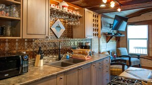 A kitchen with a microwave, sink, and wooden cabinets. There is a television mounted on the wall and a chair in the corner. - Rustic Cabin - Jetted Tubs - Wi-Fi - Wonderful Views - Grill - Firepit - Porch (Makanda)