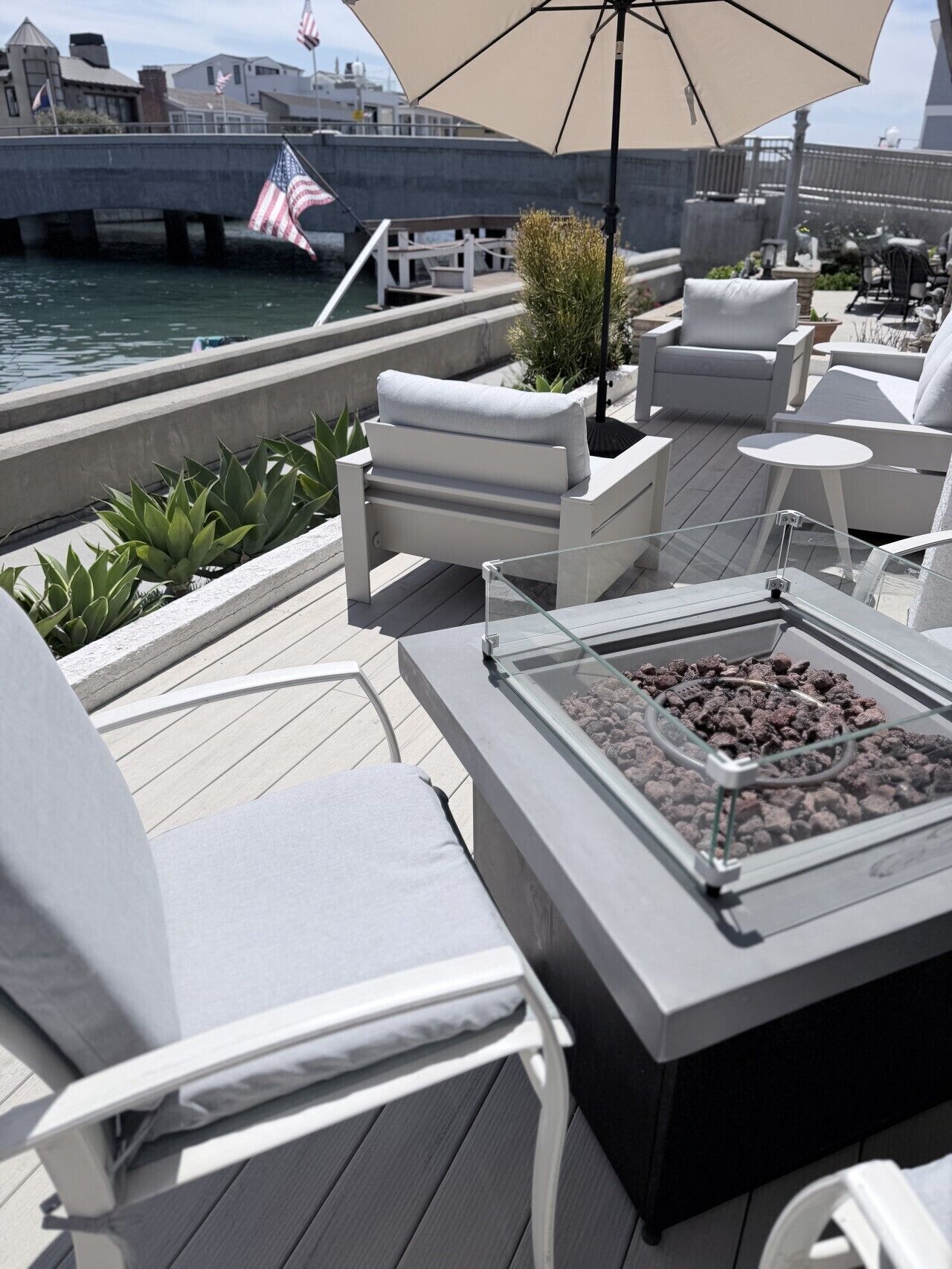 Eine Terrasse mit Sitzgelegenheiten im Freien, einem Tisch mit Glasplatte und Feuerstelle sowie Blick auf das Wasser und Flaggen.