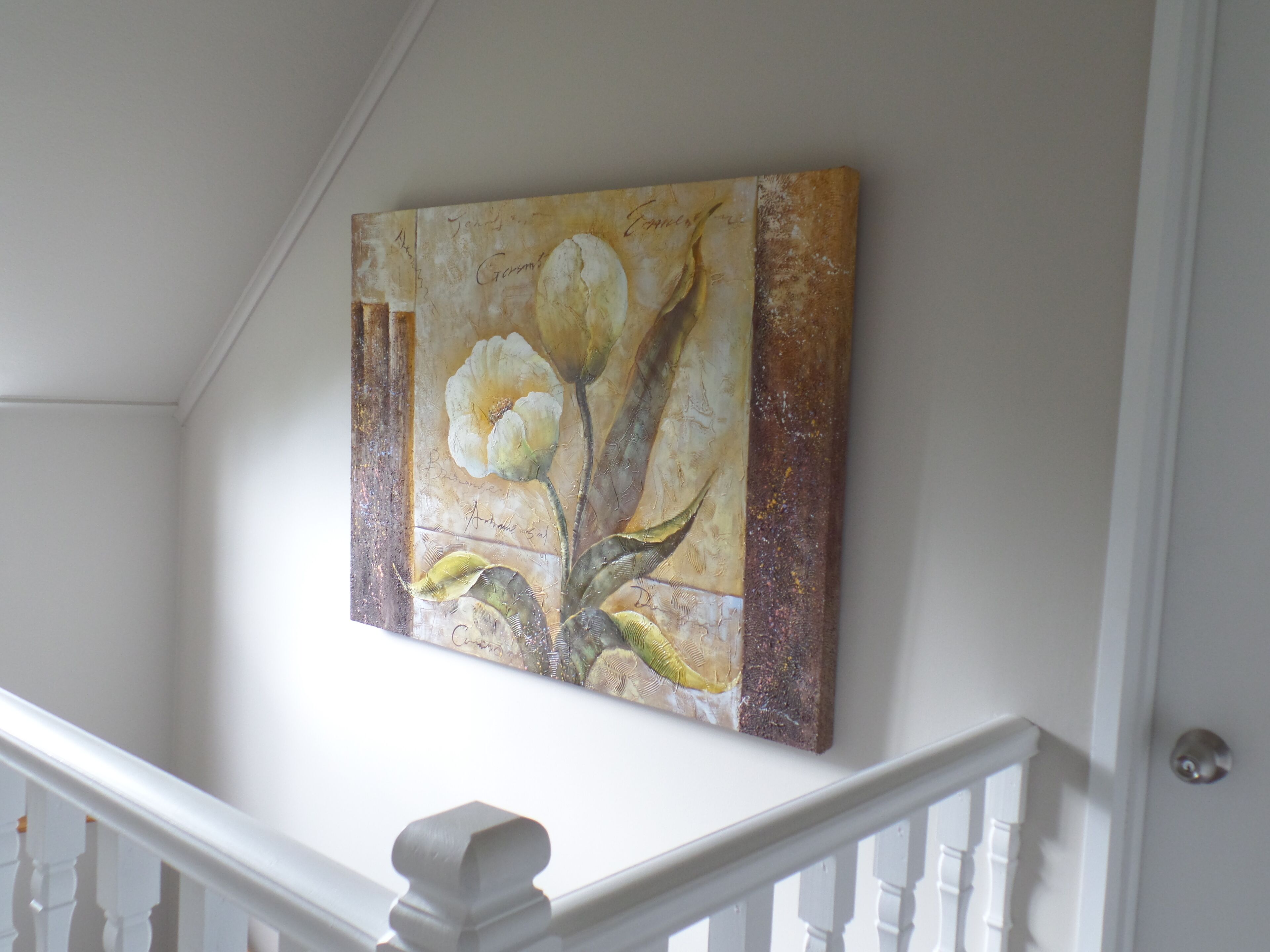 Une peinture représentant une fleur, accrochée à un mur dans une pièce aux boiseries blanches et où se trouve un escalier.