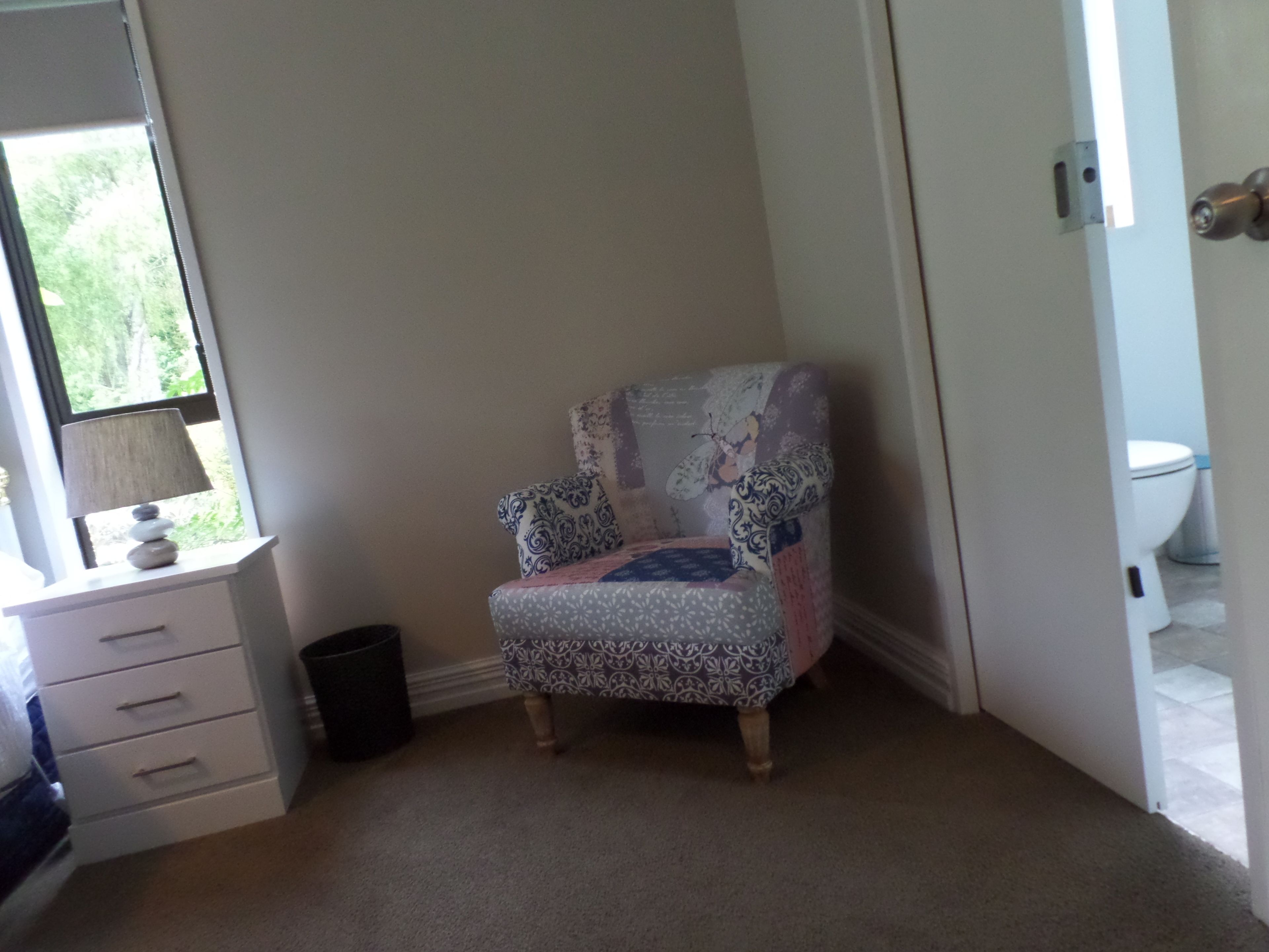 Un sillón pequeño con estampado en un rincón de una habitación, con una ventana, una puerta y una pared blanca.