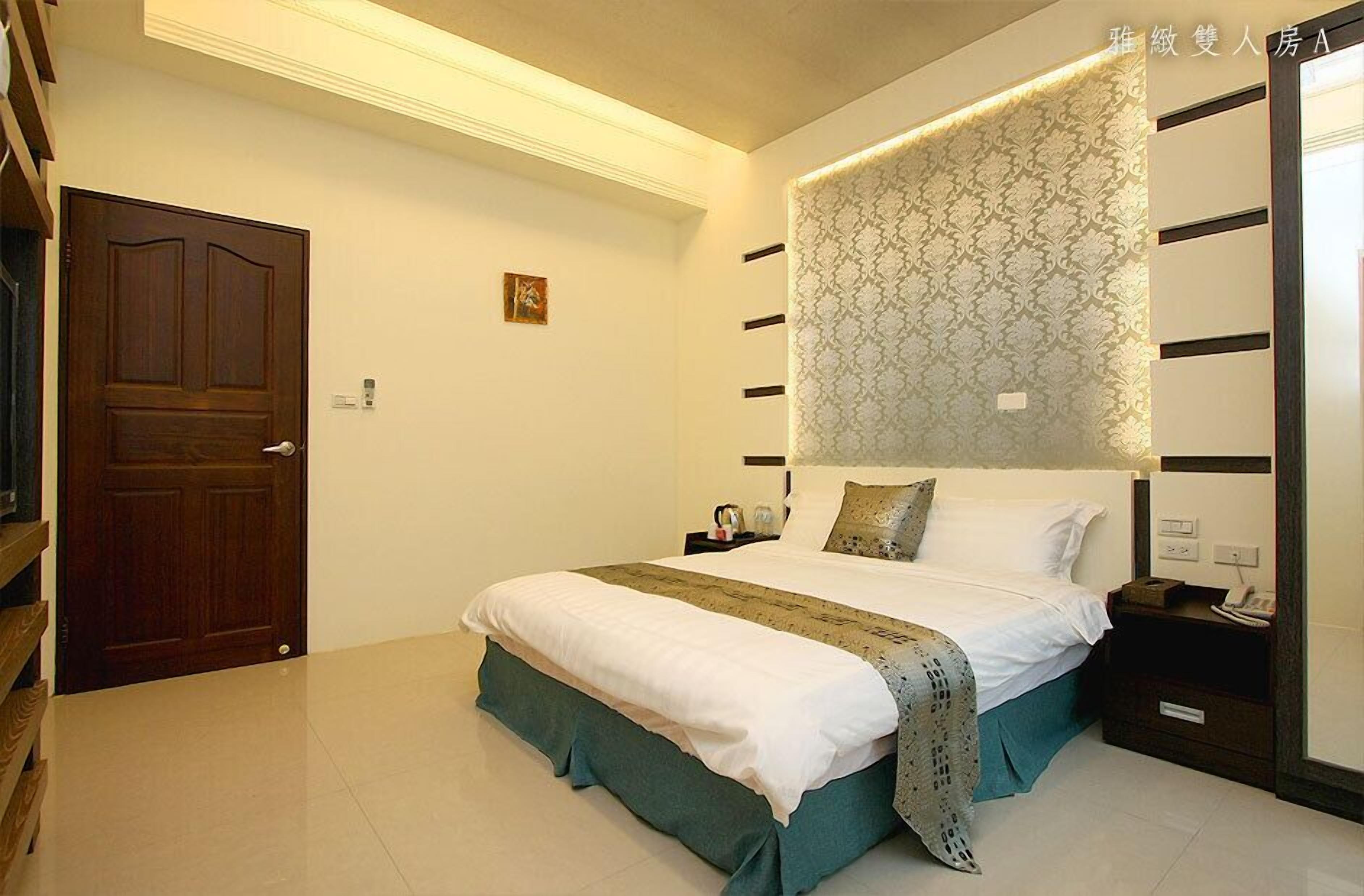 A hotel room with a large bed, a wooden door, a nightstand, and a patterned headboard.