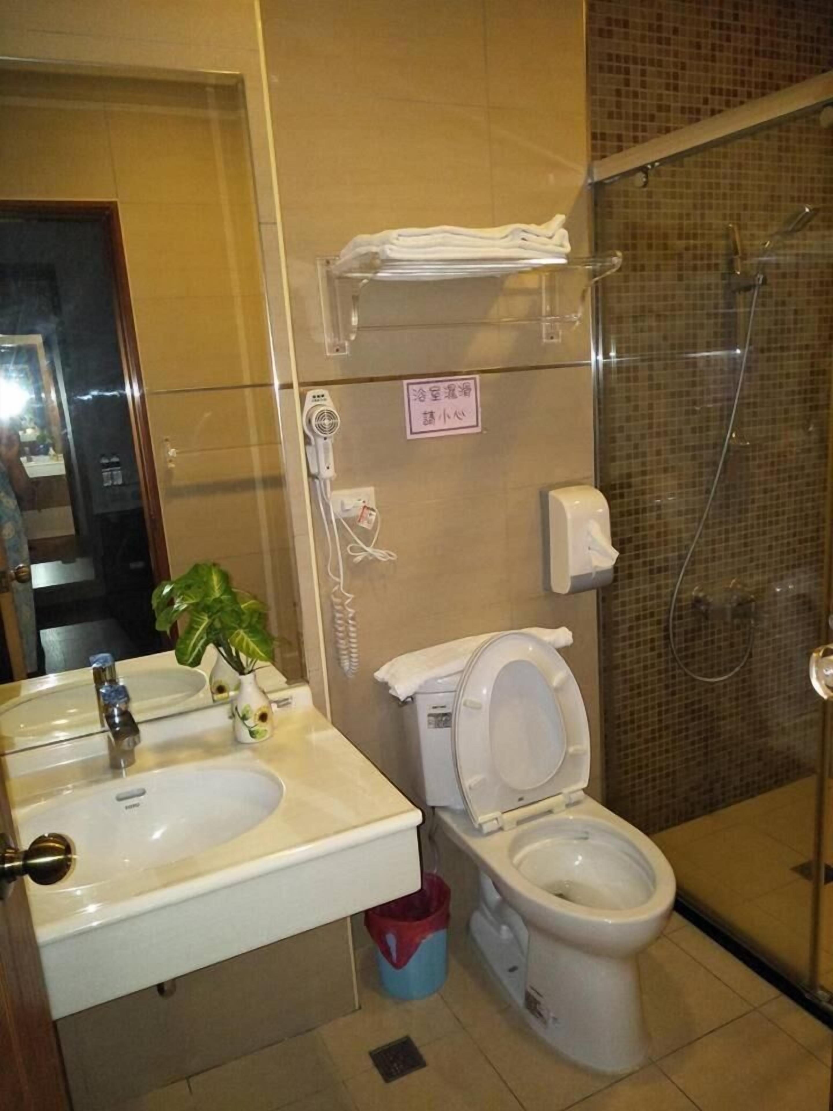 Ein Badezimmer mit Waschbecken, Toilette und Dusche. An der Wand ist ein Schild angebracht und auf der Ablage steht eine Pflanze.