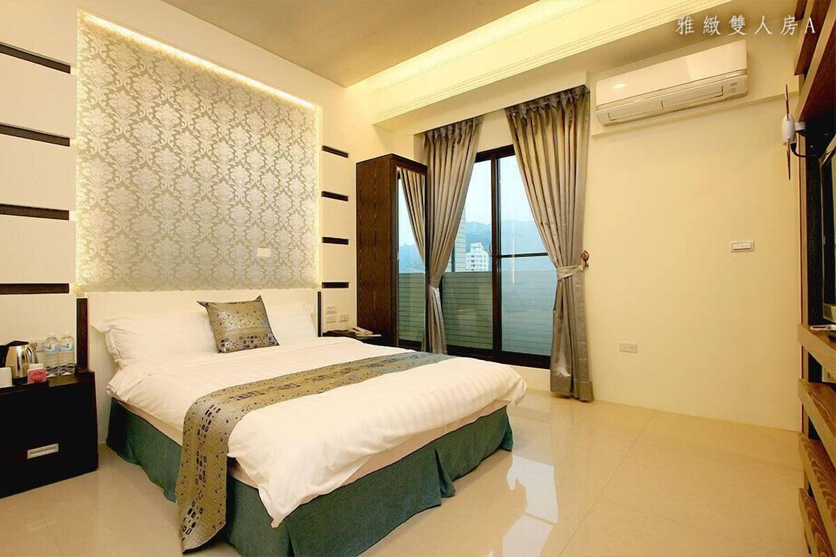 Habitación de hotel moderna con una cama grande, un panel decorativo en la pared, vista al mar y una mesita de noche de madera con artículos de cortesía.