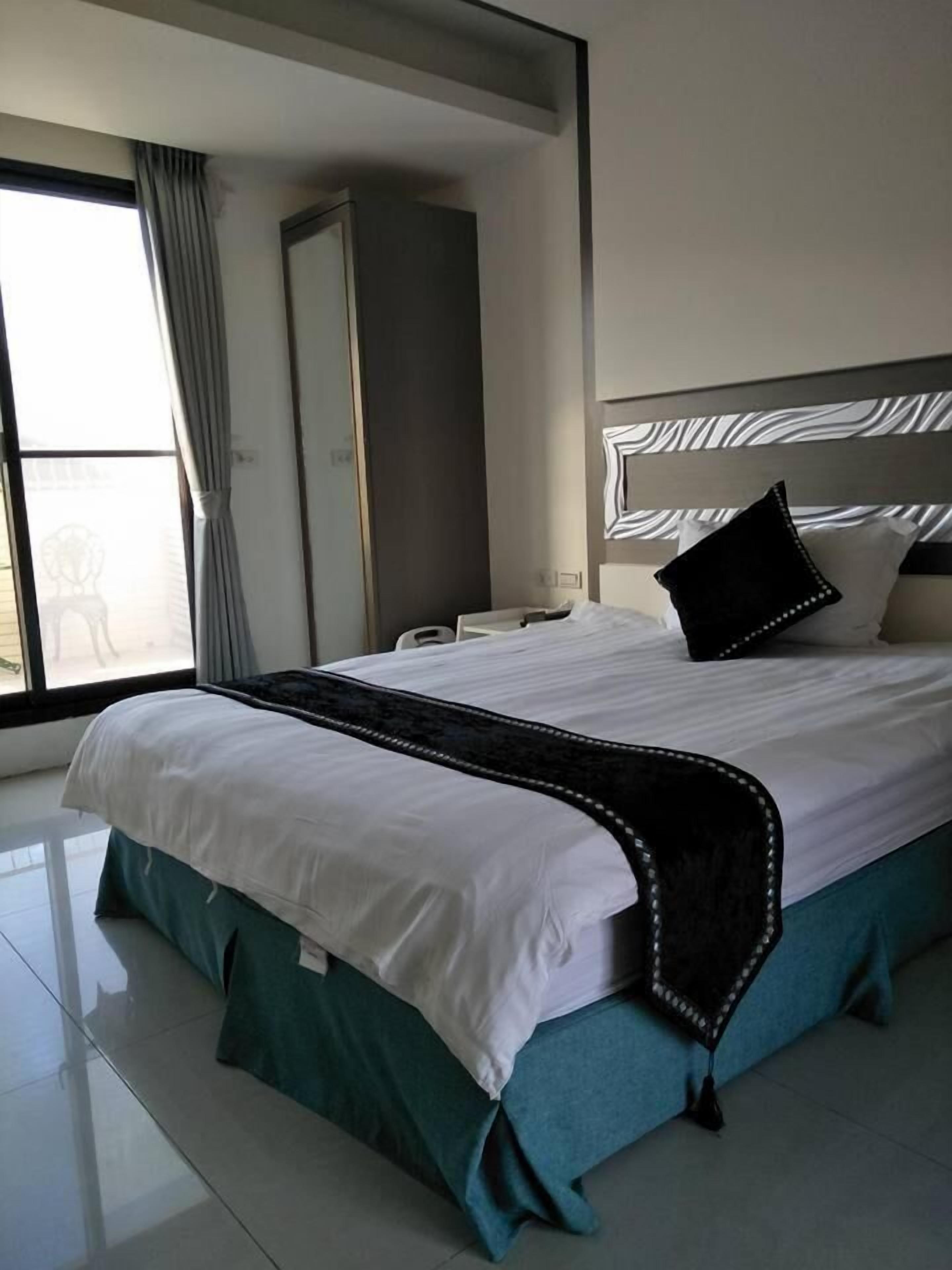 A modern hotel room with a large bed, a nightstand, a mirror, and a view of the outside through a large window.